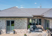 Sueddeutscher Biber Rundschnitt in silbergrau auf Walmdach-Bungalow mit Klinkerfassade mit Fokus auf die heimelige Terrasse.