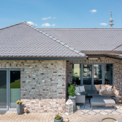 Sueddeutscher Biber Rundschnitt in silbergrau auf Walmdach-Bungalow mit Klinkerfassade mit Fokus auf die heimelige Terrasse.