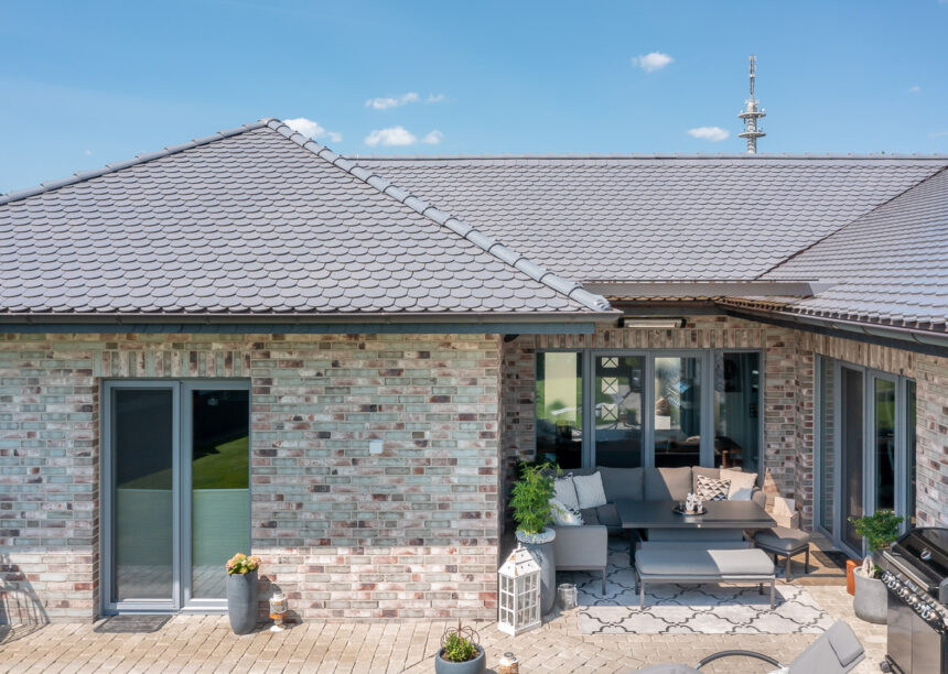 Sueddeutscher Biber Rundschnitt in silbergrau auf Walmdach-Bungalow mit Klinkerfassade mit Fokus auf die heimelige Terrasse.